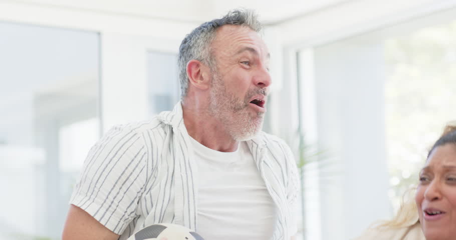 Diverse friends reacting to man holding soccer ball, high-fiving in red jersey, hugging in sunroom. Celebration, companionship, joy, teamwork, sunlit, home, candid