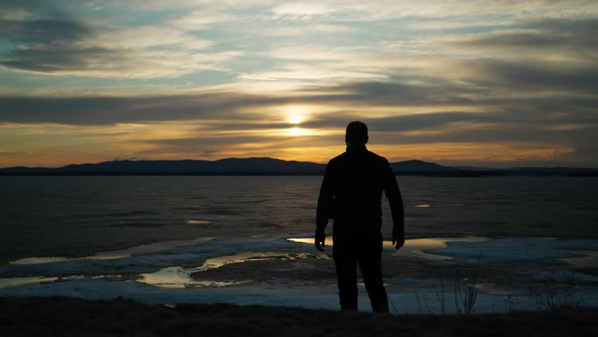 The silhouette of an enthusiastic man standing on the shore of a lake with melting ice at sunset and raising his arms to the sky. A happy person enjoys life, achievements and success. Rear view.