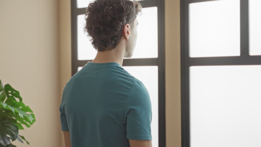 Man looking out frosted window with bare neck visible, standing near houseplant in studio, wearing teal shirt; serenity.