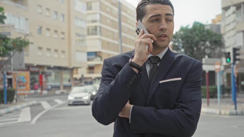 Man in navy suit and tie holding smartphone to ear and gesturing with hand on a busy street intersection, mid conversation; professional concern.
