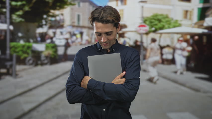 Man holding laptop with crossed arms on a street cafe with cobbles and tram tracks, looking sideways; contemplation.