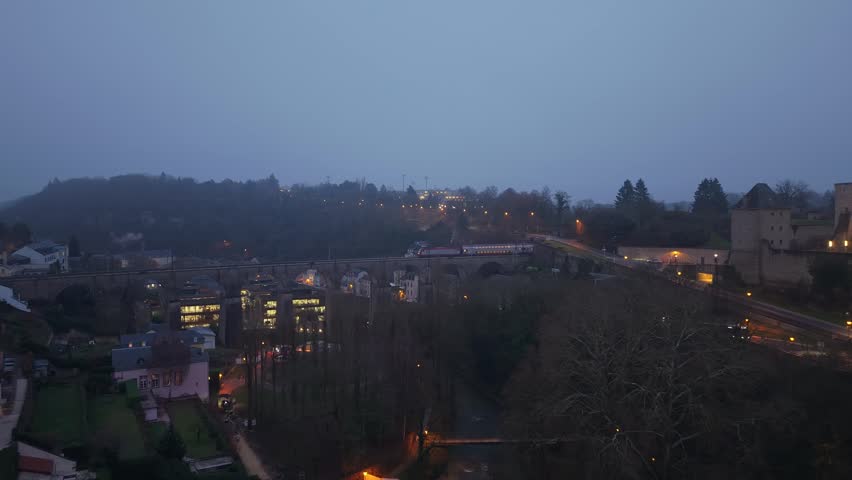 Drone reveals a train on a high winter bridge, traditional houses below, and a modern, misty skyscraper skyline.