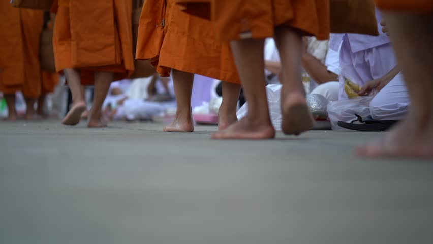 Barefoot monks in orange robes walk past kneeling people in white. The low angle highlights feet, fabric and the ritual path.
