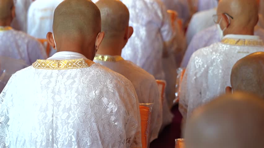 A rear perspective shows white clad ordinands seated in rows inside the temple holding orange robes with shaved heads and folded hands in a soft blurred ritual setting.