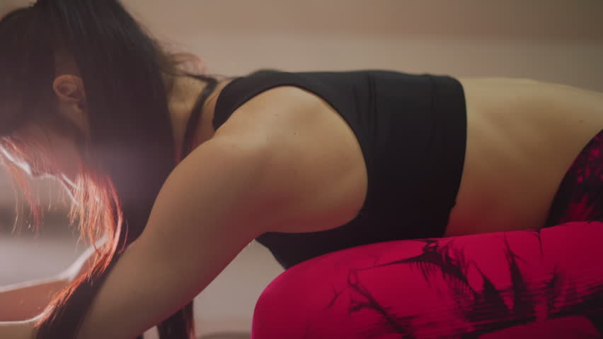 asian woman stretching on mat in studio, performing child pose for deep spinal release, red leggings and black sports bra lit by warm window light, slow breath and mindful relaxation, yoga student