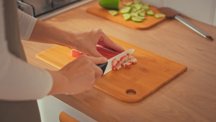 A woman, like a chef, deftly slices bright crab sticks on a kitchen counter, mixing them with fresh vegetables and herbs. The careful cooking process showcases home kitchen food preparation