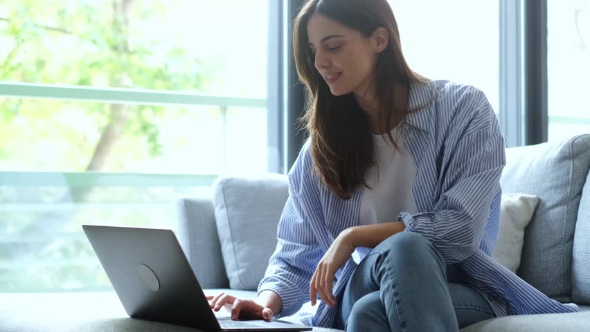 A happy young woman is smiling and focused while using her laptop. She sits comfortably on a modern sofa at home, perhaps engaged in remote work, online learning, or casual browsing.