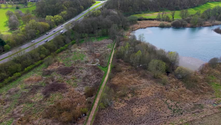 Panshanger Park aerial pan over lakeside wetlands and the A414 highway, showing woodland edges and open parkland in fresh spring colours, Hertfordshire, England.