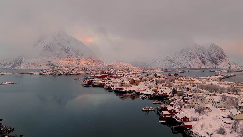 A breathtaking drone shot of the snowcovered fishing village of Reine in Lofoten, Norway, featuring traditional red cabins and dramatic mountain peaks under a soft winter glow.