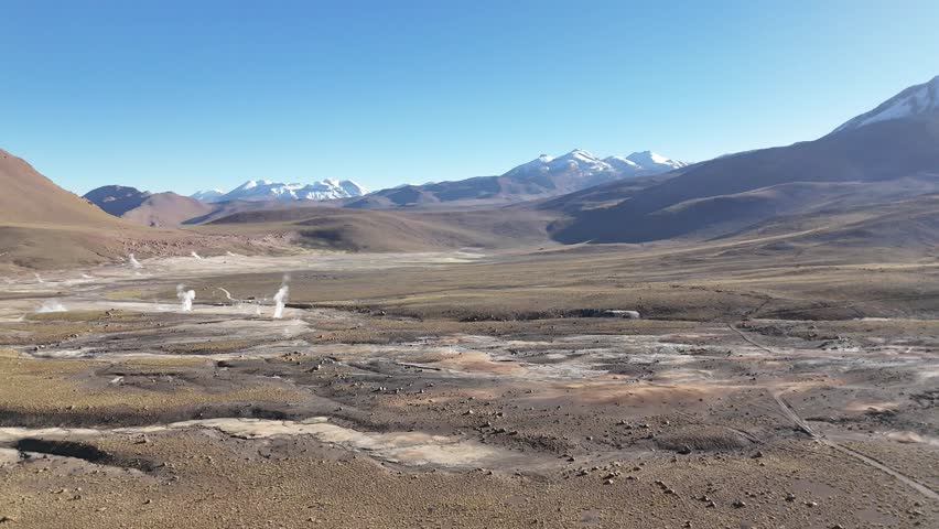 Cinematic aerial flying forward over steaming geothermal vents and colorful mineral runoff at El Tatio, revealing the vast volcanic plateau of the Chilean Altiplano