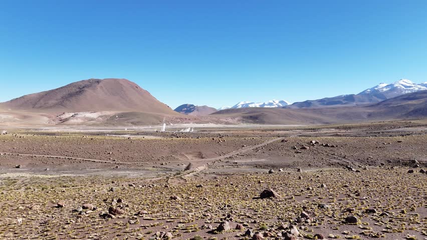 Cinematic aerial flying forward over steaming geothermal vents and colorful mineral runoff at El Tatio, revealing the vast volcanic plateau of the Chilean Altiplano