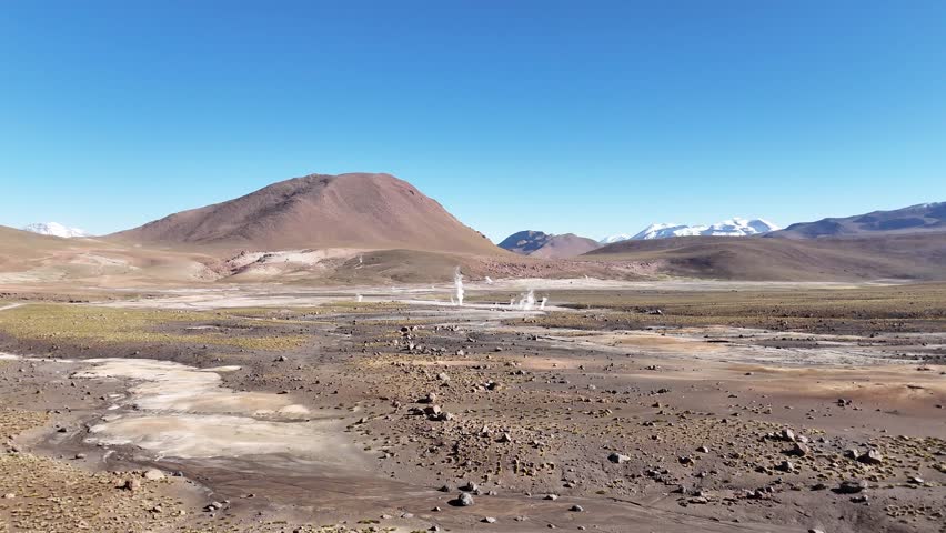 Cinematic aerial flying forward over steaming geothermal vents and colorful mineral runoff at El Tatio, revealing the vast volcanic plateau of the Chilean Altiplano