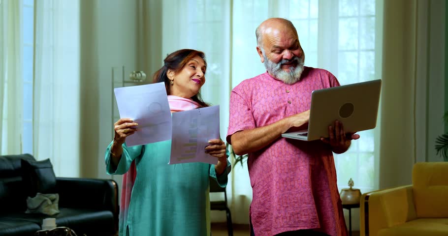 Senior Indian couple standing in a modern living room using a laptop and documents to manage their budget, then smiling and showing a thumbs up to the camera