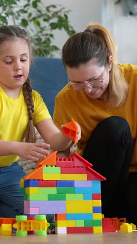 A woman and a girl build a colorful house with building blocks. They focus on the structure made of various blocks while sitting on a patterned mat in a bright room.
