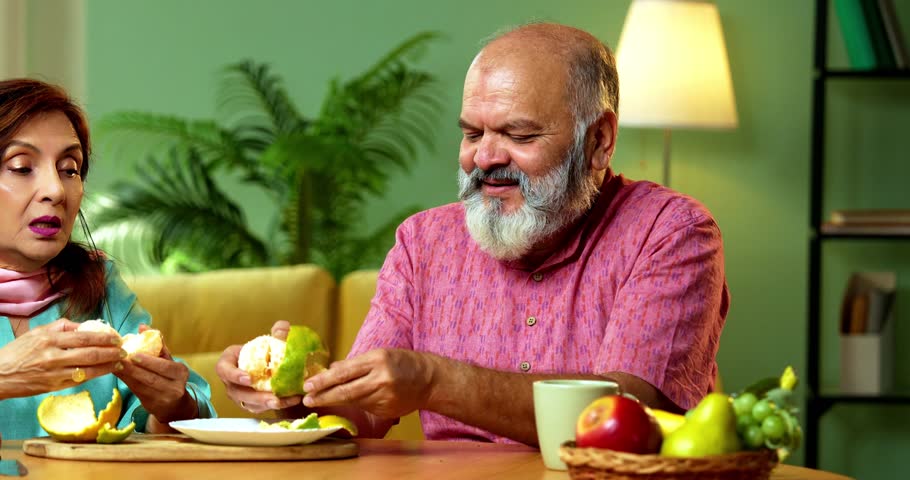 Senior Indian couple sitting together in their modern home while peeling fresh orange fruit and enjoying the juicy slices with smiles and conversation