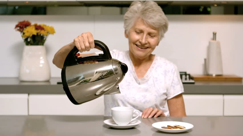 Retired Caucasian woman pouring boiling water from kettle into cup in kitchen in slow motion