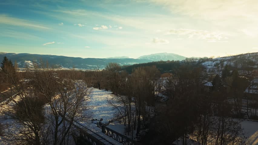 Drone ascending over snow covered train tracks in Enveig with village and mountains in background during golden hour.