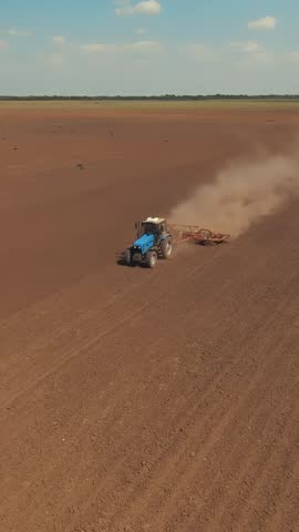A blue tractor is working in a large field, plowing the brown soil. Dust rises from the tractor as it moves across the expansive agricultural land.