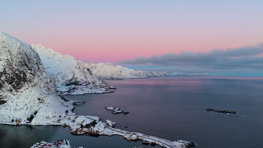 Breathtaking aerial view of the Lofoten islands in Norway during a pink winter sunset, featuring snowcapped mountains and a calm arctic fjord.