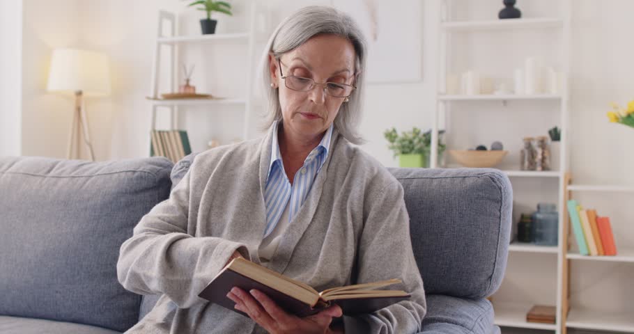 Senior peaceful retired woman book reading, elder positive mature grey haired lady resting at home, intelligent interesting wearing glasses to enjoy novel having rest on comfortable home couch alone