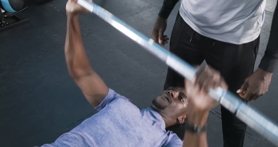 After inhaling African American man bench pressing barbell with spotter at gym for strength. Strength, fitness, training, athletic, determination, motivation, power