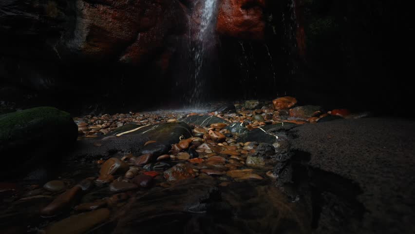 Tilt up shot from rocky stream bed to waterfall at Dicido Beach revealing wet cliffs, flowing water and vegetation above