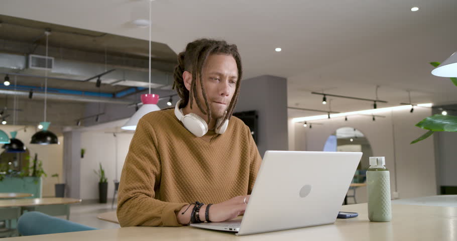 African American man typing on laptop at coworking table, glancing, reaching green bottle, drinking. Focus, productivity, hydration, workspace, modern, openplan, plants