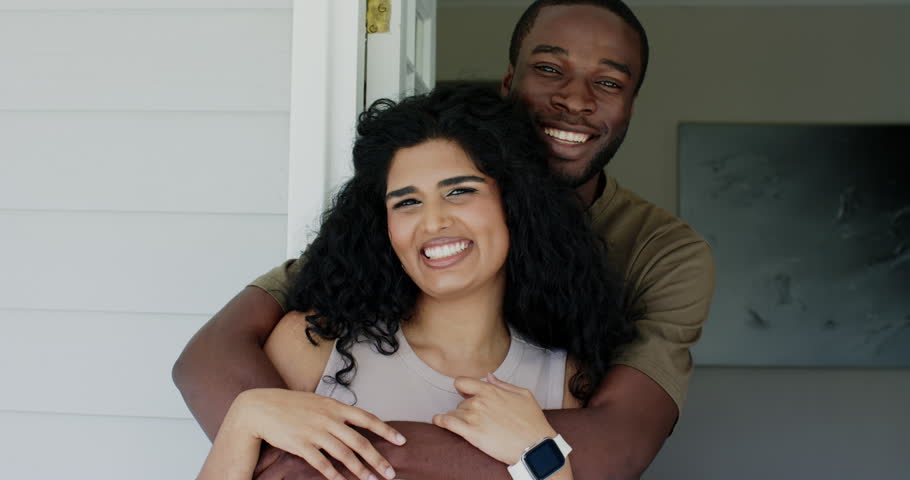 Diverse couple in doorway, man wrapping arms, woman holding cushion, smartwatch, laughing, posing. Porch, threshold, home, warmth, affection, connection, softlight