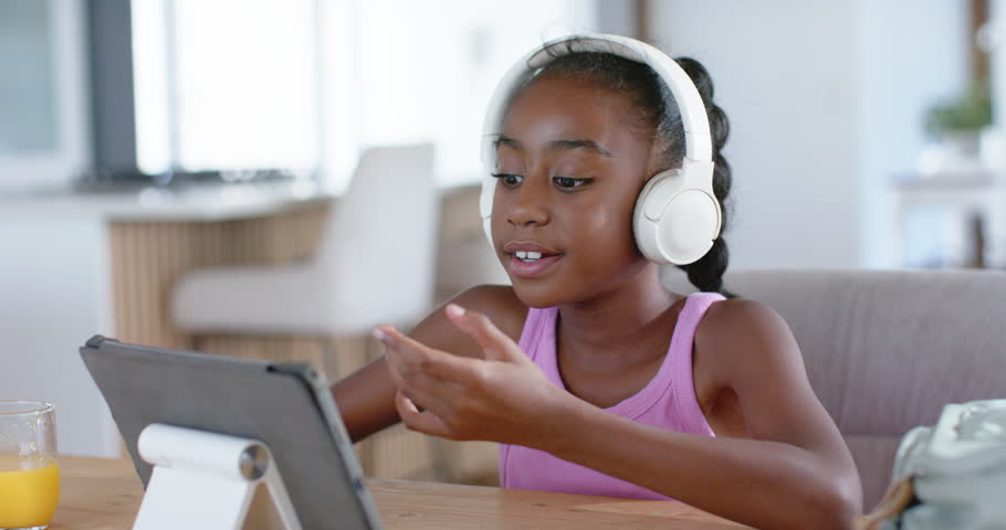 African-American girl at home with headset responding to teacher prompt, leaning in, tapping tablet. Child, onlineclass, education, device, headphones, gestures, dining