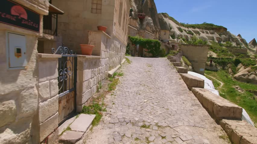 Footage capturing a picturesque, narrow cobblestone street winding its way through the unique landscape of Cappadocia, Turkey. Traditional stone and cave houses, built into the fascinating rock format