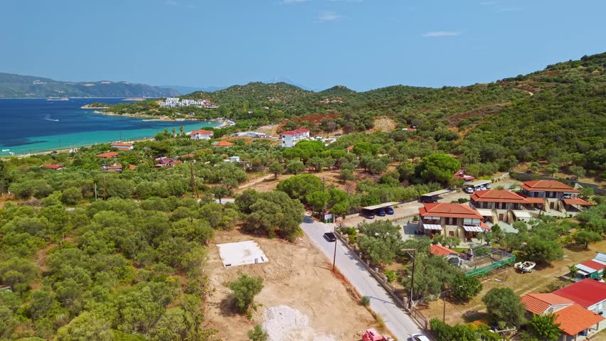High angle aerial drone shot of the turquoise Aegean Sea coastline with white houses and green trees near Paralia Glastri beach, Greece