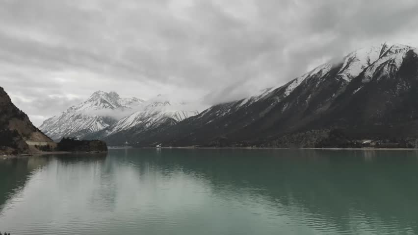 Dramatic horizontal timelapse of Ranwu Lake in Tibet. Clouds move over snow-capped peaks perfectly reflected in the turquoise mountain lake.