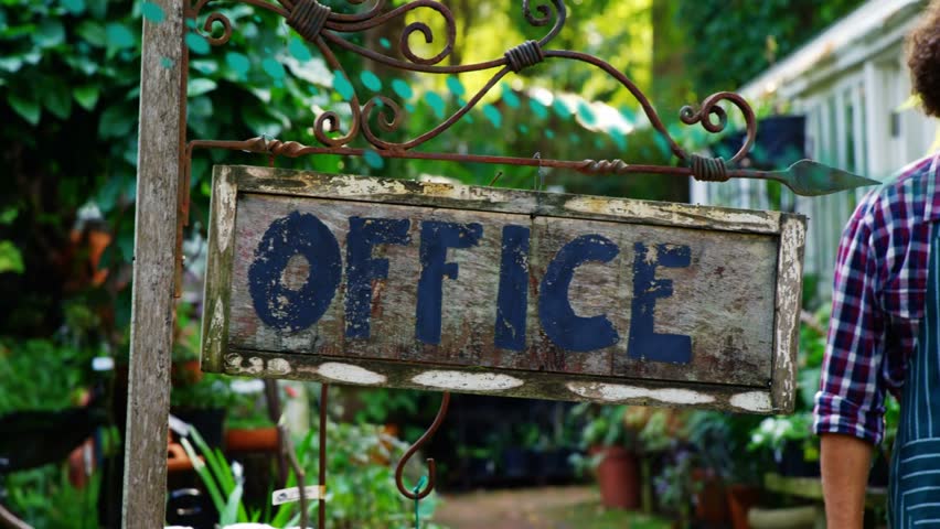 Shop owner entering with Open sign, adjusting OFFICE bracket and signaling plant nursery opening. Chalkboard, greenery, storefront, greenhouse, rustic, sunlight, welcoming