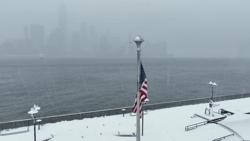 American flag waving in strong winter wind along snowy waterfront promenade in Jersey City with Manhattan skyline fading into fog across Hudson River during cold snowstorm aerial orbit view.