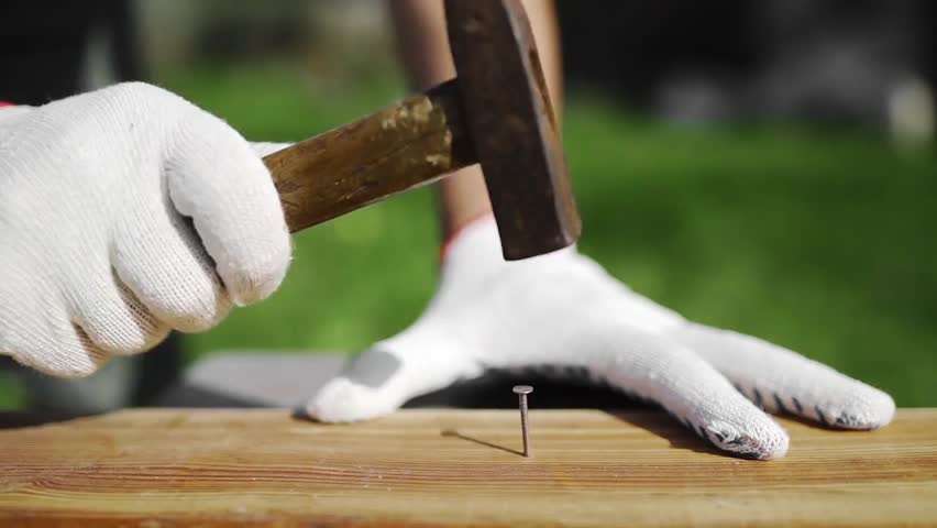 Man wearing working gloves hammering a nail outdoors, repairing wooden furniture in backyard DIY work, showing home repair and restoration.