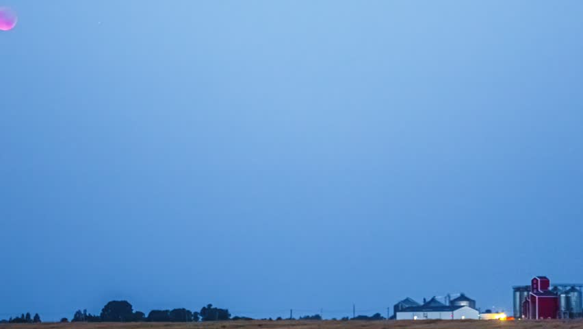 Moon sets over a farm field and silo beneath a clear blue twilight sky in timelapse