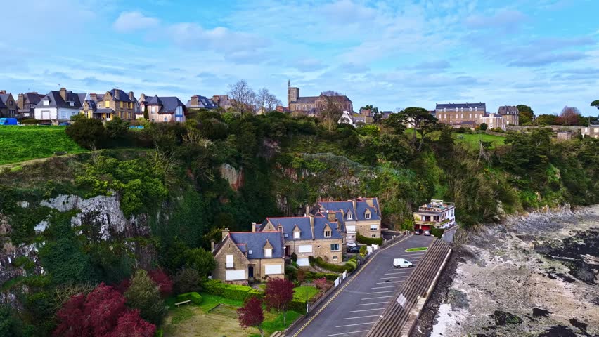Drone rises over Cancale, Brittany, revealing stone houses, rocky shoreline, and Saint-Méen Cathedral towering above green cliffs on a sunny day