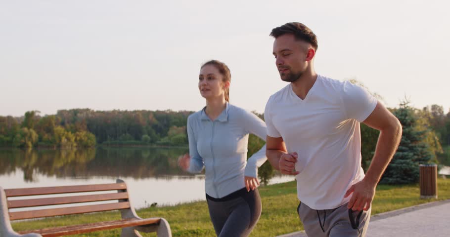 Morning healthy run of fit couple jogging marathon on sidewalk at summer lake. Active happy woman and man in sportswear training outdoors, young two athletes running together on trail in park