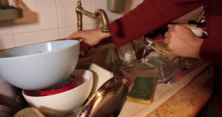 Male hands washing glassware with a sponge under running water in a messy kitchen sink