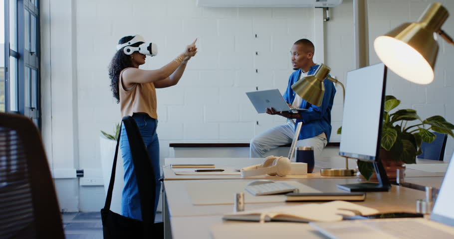 Colleagues at desk, woman initiating VR gestures with headset, guiding man using laptop. Female, male, office, openplan, technology, virtual, collaboration