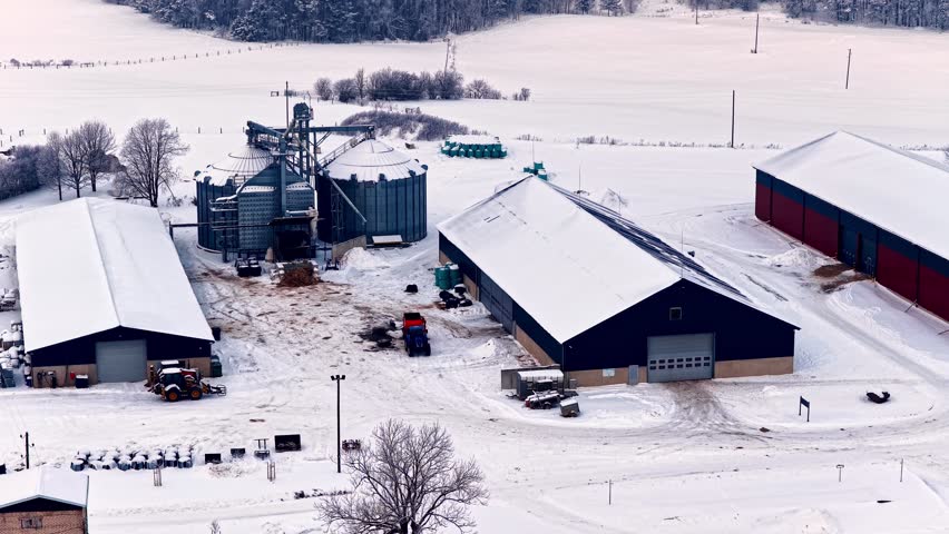 Snowy farm buildings with grain silos and barns in winter countryside, aerial pullback