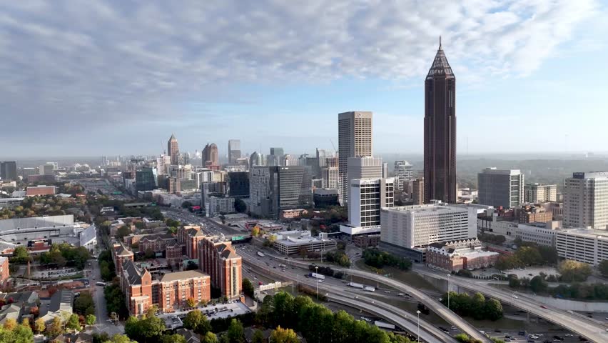 A sweeping aerial drone shot capturing the sprawling downtown Atlanta skyline under a clear blue sky. The footage highlights iconic skyscrapers like the Westin Peachtree Plaza and buildings in the distance, while the foreground showcases the bustling Downtown Connector with heavy traffic flow. This high-angle perspective perfectly illustrates the urban density, modern architecture, and the high-energy transportation network of Georgia’s capital city.