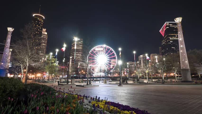 A breathtaking aerial drone shot of SkyView Atlanta, the iconic Ferris wheel located in Centennial Olympic Park. The wheel is brilliantly illuminated with vibrant LED lights, spinning slowly against the backdrop of the glowing Atlanta, Georgia skyline at night. This cinematic perspective highlights the city
