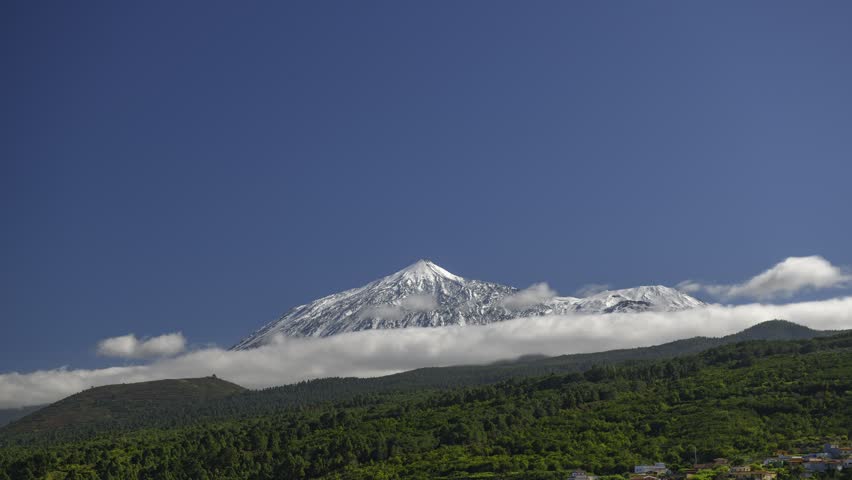4K time-lapse video showing white clouds dramatically flowing and wrapping around the snow-covered summit of Mount Teide, the highest peak in Spain. Filmed in Teide National Park on Tenerife, Canary Islands, under a clear deep blue sky. The dynamic movement of clouds creates a powerful and serene natural spectacle. Ideal for nature documentaries, travel content, cinematic backgrounds and atmospheric video projects
