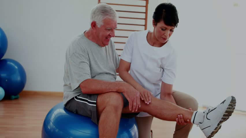 Therapist cueing and holding leg for therapy, lifting, guiding on blue ball with DNA helix overlay. Rehabilitation, clinician, patient, mobility, extension, motion, stability