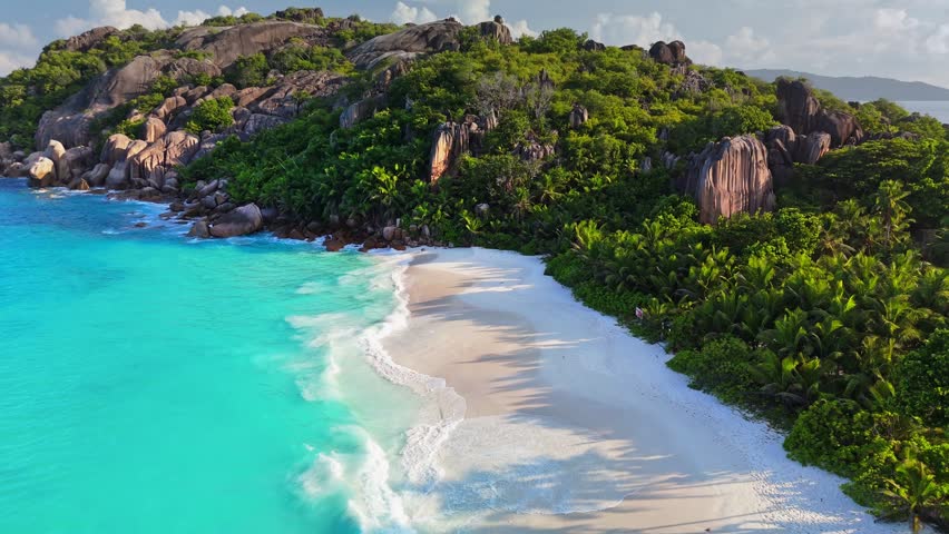 Turquoise ocean waves wash the white sand beach in the Seychelles. Island aerial captures jungle trees and granite boulders bordering the coast of the Seychelles