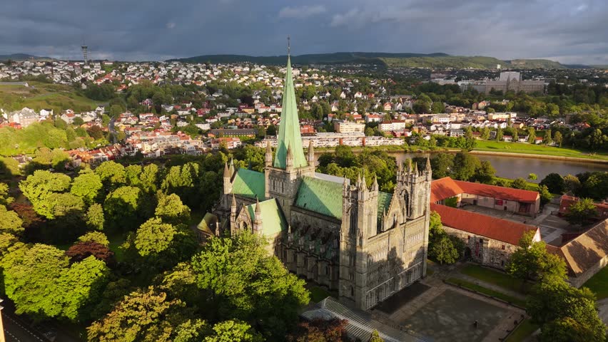 Panoramic flight aerial of the gothic Nidaros Cathedral standing among green trees in Trondheim, Norway. City buildings surround the historic Nidaros Cathedral of Norway