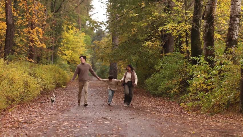 Happy family, man and woman young adults, little boy, casual clothes, running holding hands with small dog on forest path, front view, autumn trees, outdoor activity, parenting, togetherness.