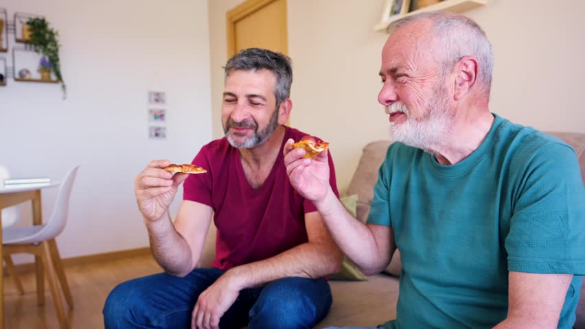 Happy mature gay couple enjoying pizza while sitting on the sofa at home. Two senior men watching a sports match together at home