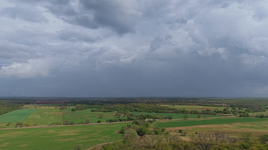 Aerial push forward and tilt up over rural farmland with a road and bridge as a storm builds in the distance under heavy clouds.